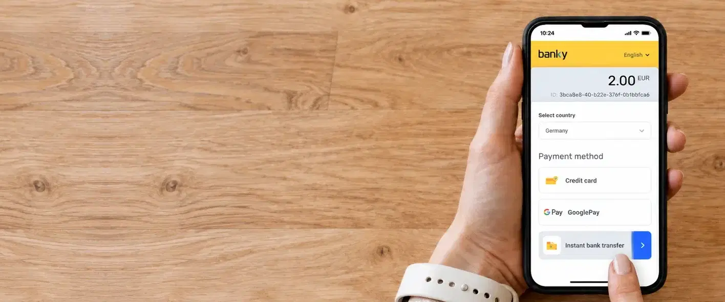 Seen from above: two hands hold a phone at the right of a pale wooden table, the Banky checkout open on it with a country chosen and a finger pressing instant bank transfer in the list of payment methods.