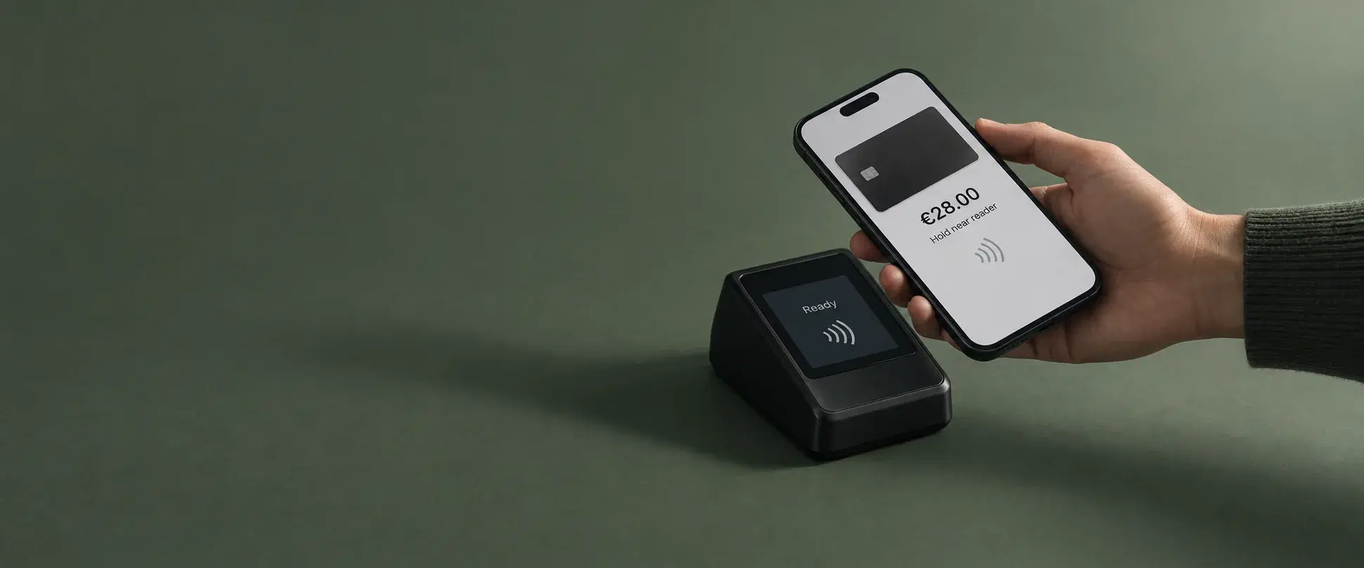 A hand holds a phone over a small black contactless terminal on a deep green surface; the phone shows a plain dark card, the amount 28.00 euros and the words hold near reader, and the terminal reads ready.