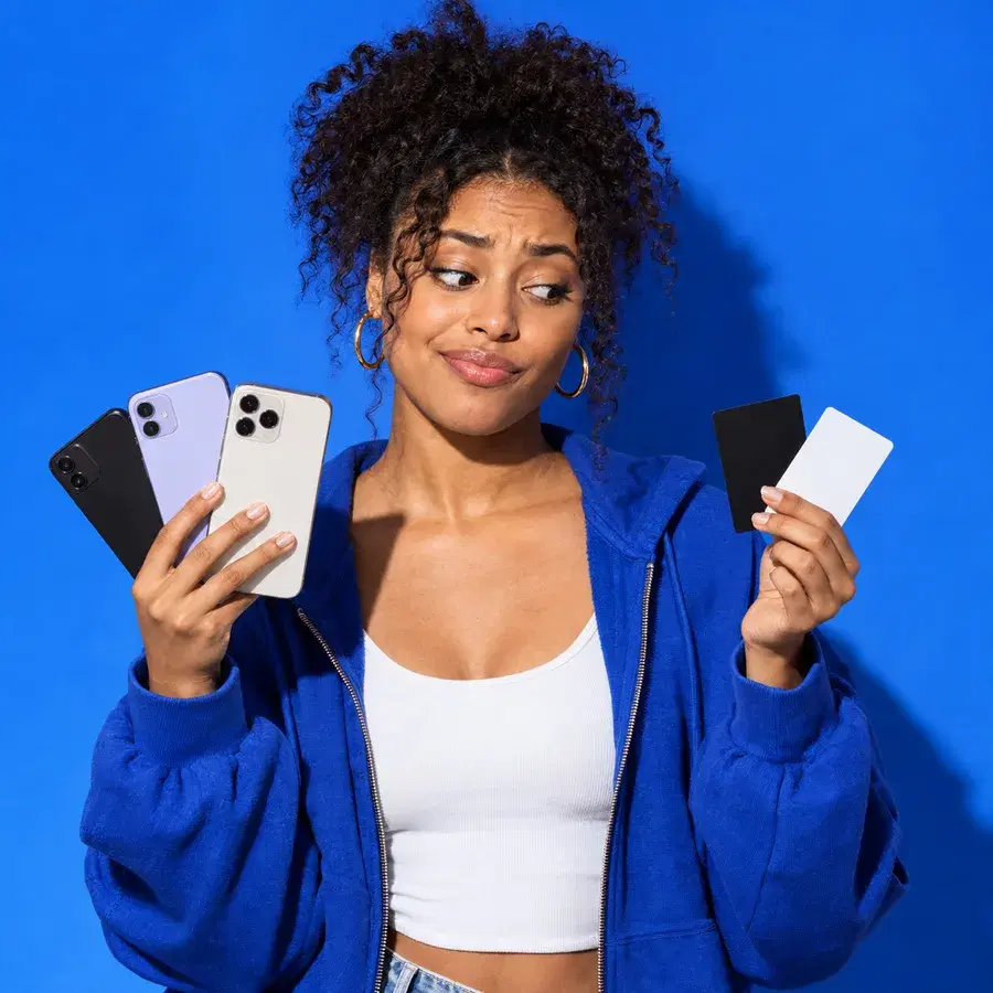 A young woman against an electric-blue backdrop, three phones fanned out in one hand and two blank payment cards in the other, looking at them with amused exasperation.