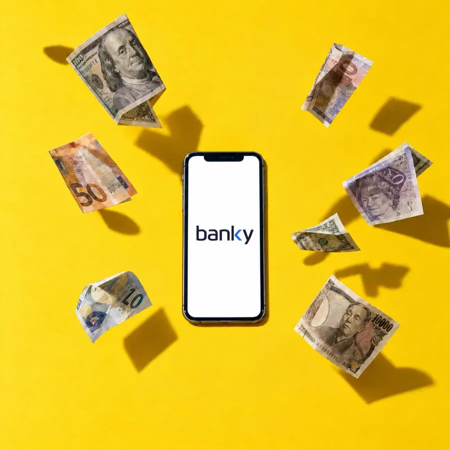 A phone lying on a bright yellow surface seen from above with the Banky mark on its screen, banknotes in six currencies floating around it and casting hard shadows.